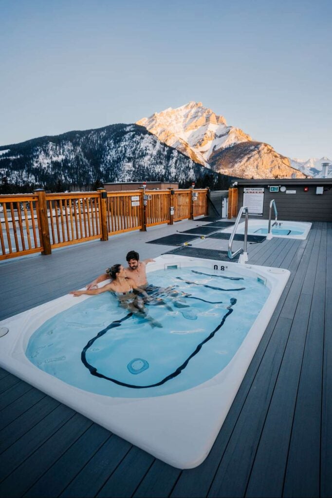 a couple relax in the rooftop hot pools on top of Basecamp Suites Banff.