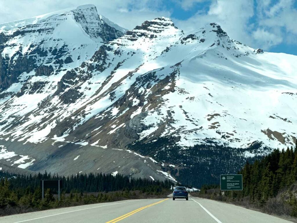 a single car is dwarfed by a snow capped mountain on the Icefields Parkway near the Glacier Discovery Centre.