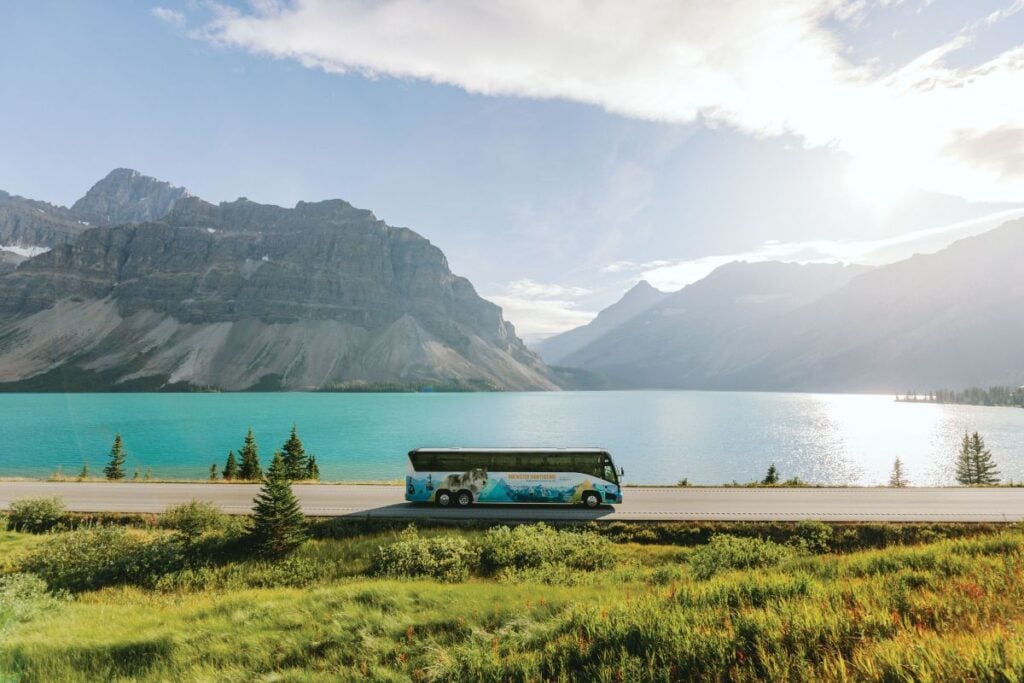 The Brewster Express airport shuttle bus drives next to a beautiful lake and a backdrop of Rocky Mountains.