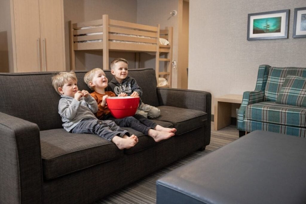 Three kids sit on the couch with a bowl of popcorn in a family-friendly hotel room at the Mountaineer Lodge in Lake Louise, Canada.