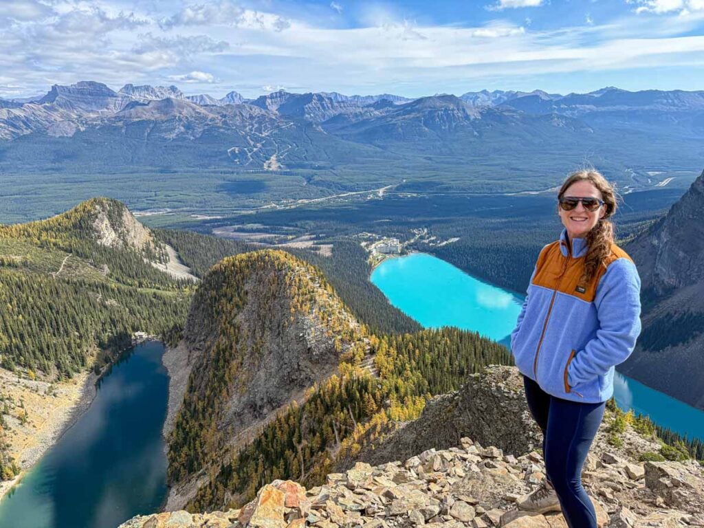 Celine Brewer, host of the Banff Travel Podcast, stands at the summit of the Devil's Thumb Trail overlooking Lake Louise and Lake Agnes.