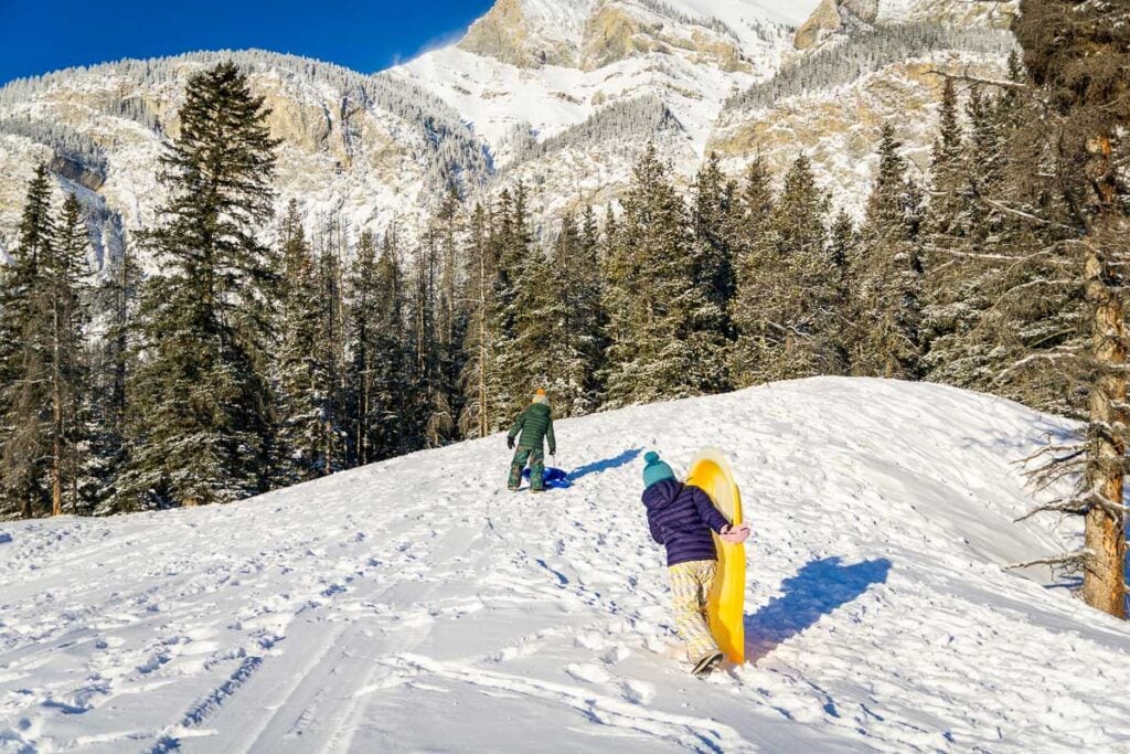 Two Brewer kids carrying sleds up a snowy hill at Cascade Ponds in Banff.